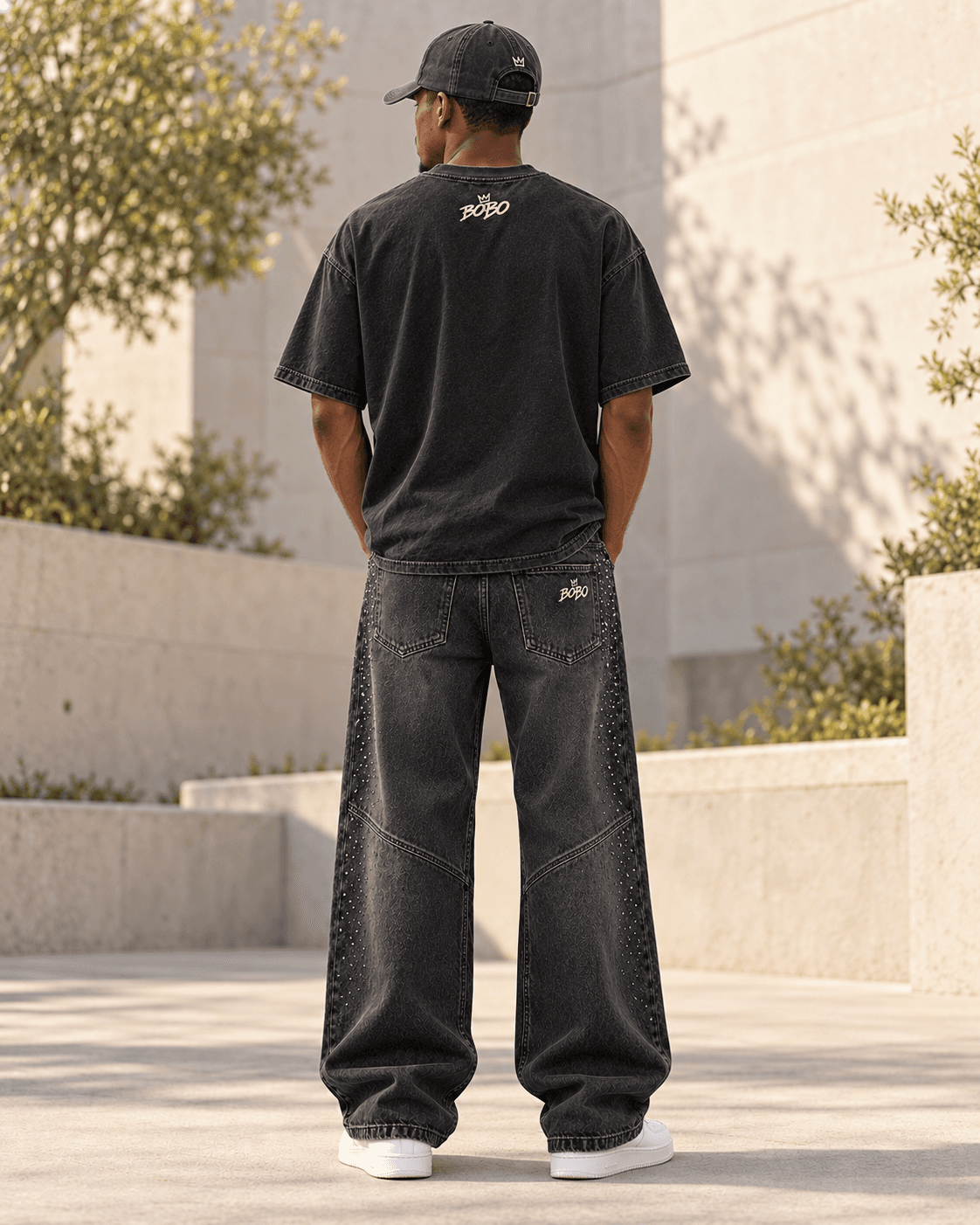 Rear full-body view of the black BOBO Crown Graphic Tee, crystal charcoal denim and grey crown cap in the same sunlit courtyard
