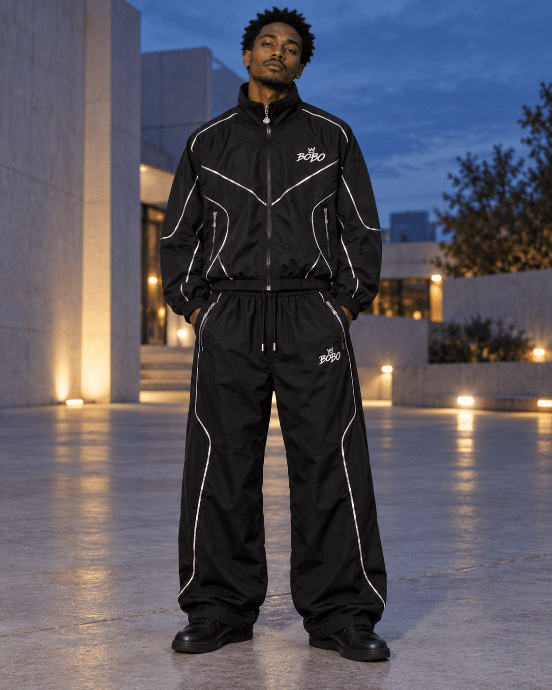 Full-body model wearing a black BOBO Reflective Tech Track Set with silver seam lines and black sneakers in a plaza at blue hour