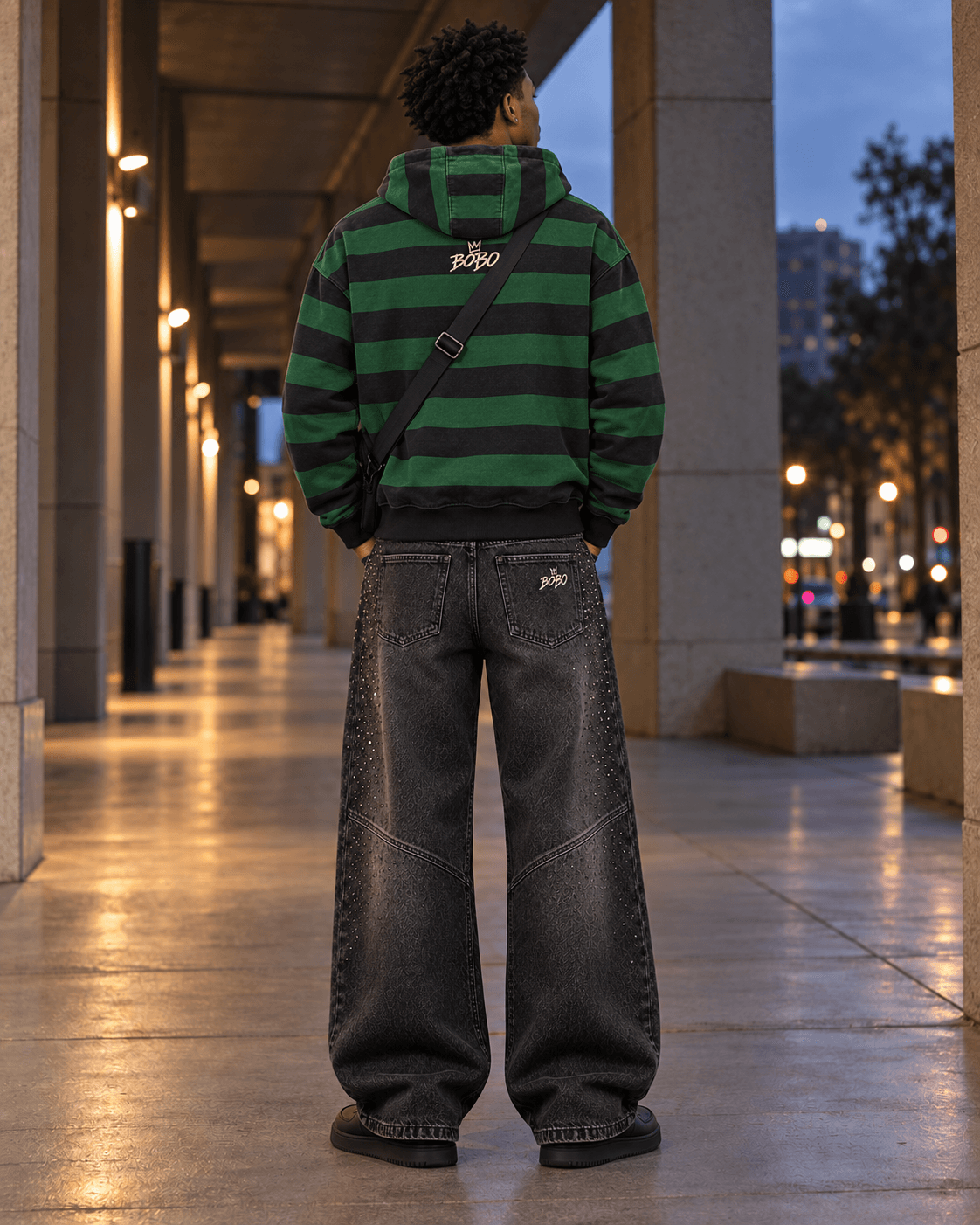 Rear full-body view of the BOBO striped zip hoodie and crystal charcoal denim with a crossbody strap under the same city walkway